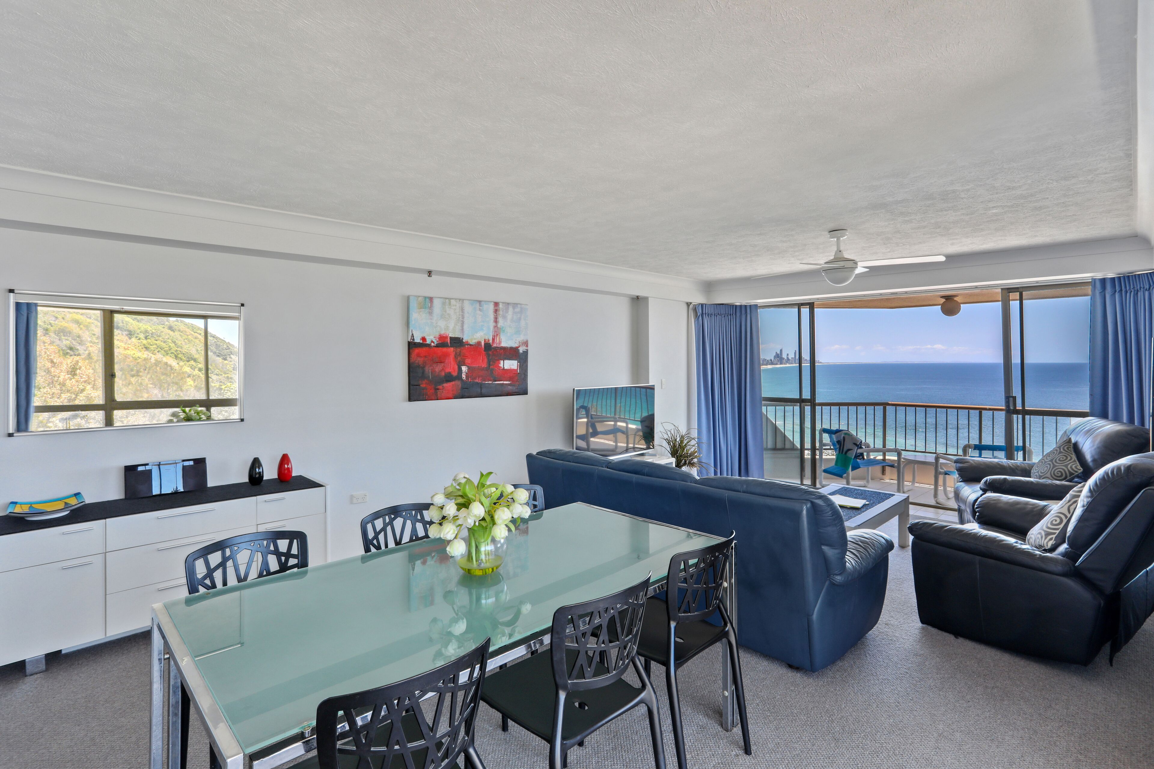 Uma sala de estar moderna com uma mesa de jantar de vidro, cadeiras pretas, um sofá azul e vista para o oceano.