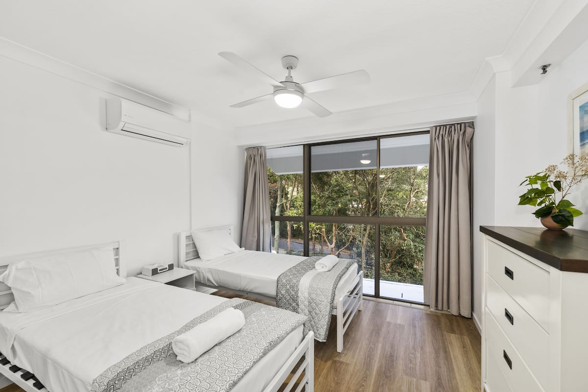 A modern hotel room with two beds, a ceiling fan, an air conditioning unit, a large window with curtains, and a plant on the dresser.