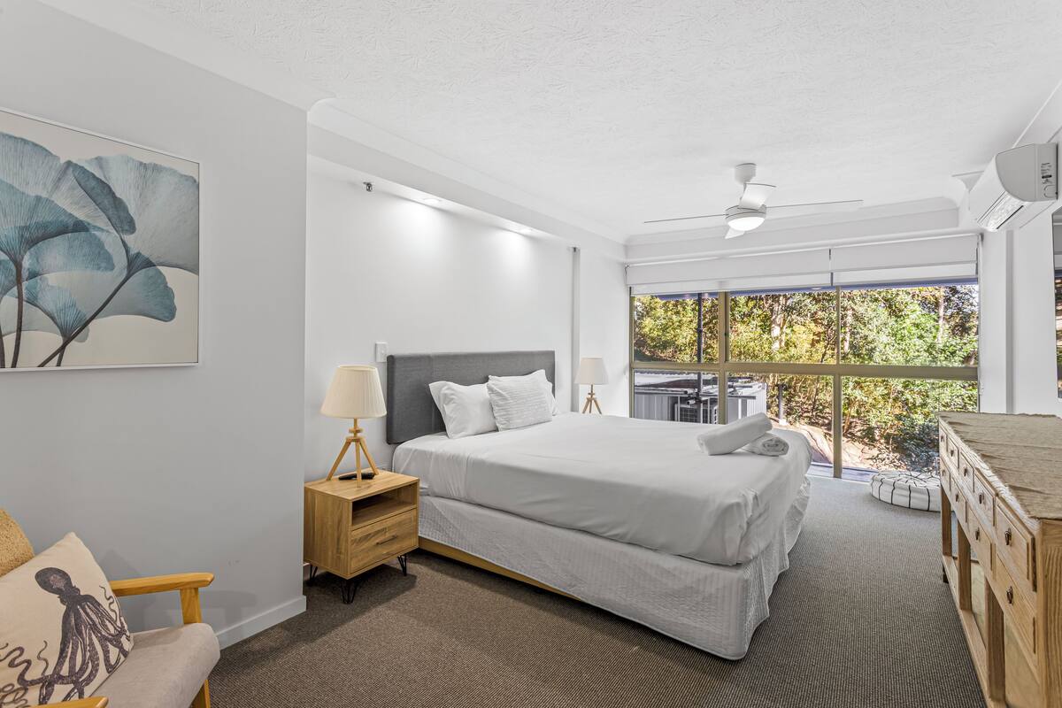 A modern bedroom with a large bed, bedside lamps, a nightstand, a wooden dresser, and a large window with a view of greenery.