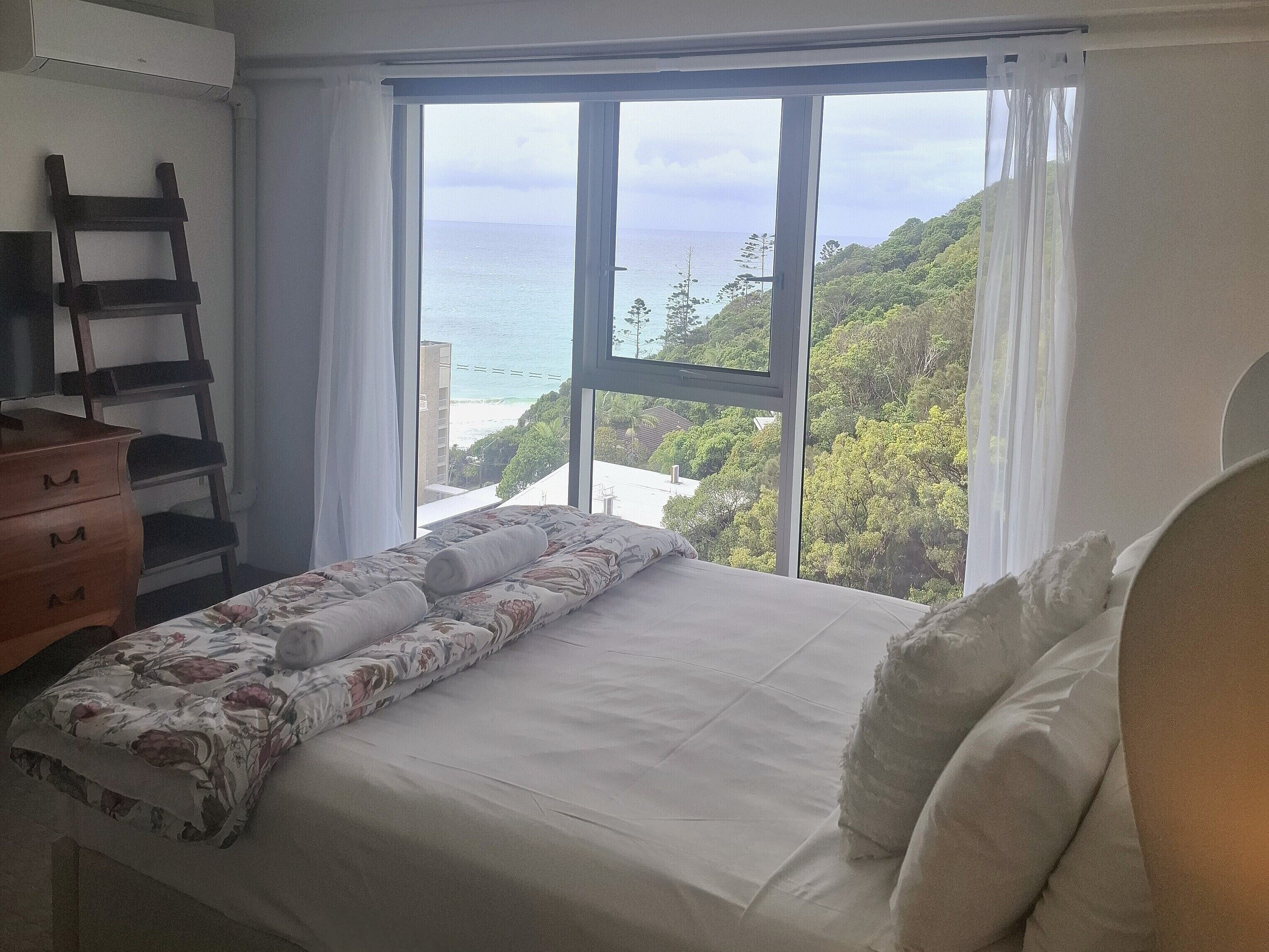 Una camera da letto con un letto, una cassettiera, una sedia e una finestra vista mare e vegetazione.