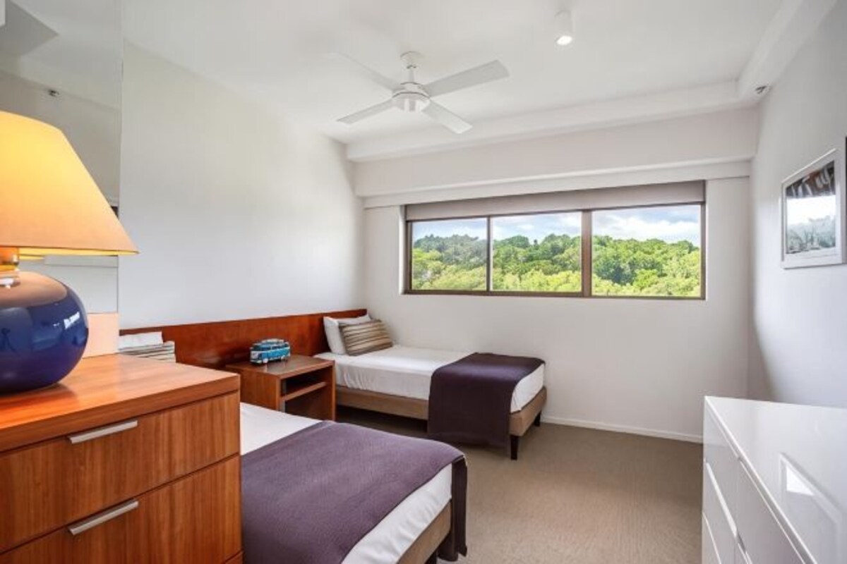 A hotel room with a bed, a desk, a chair, a nightstand, a lamp, a ceiling fan, and a window with a view of greenery.