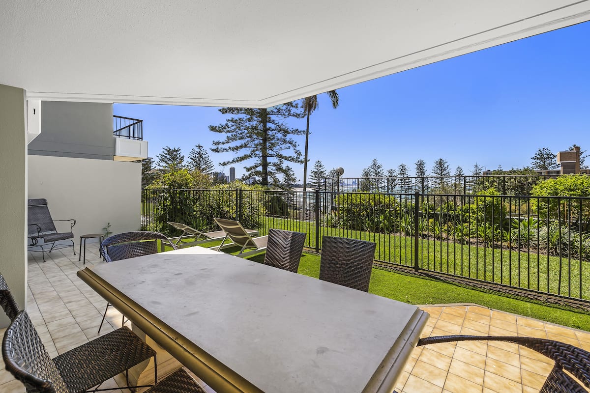 A balcony with a table and chairs, a view of a well-maintained garden, and a clear blue sky.