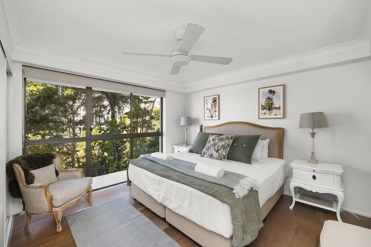 A bedroom with a large bed, a chair, a nightstand, a ceiling fan, and a window with a view of trees.