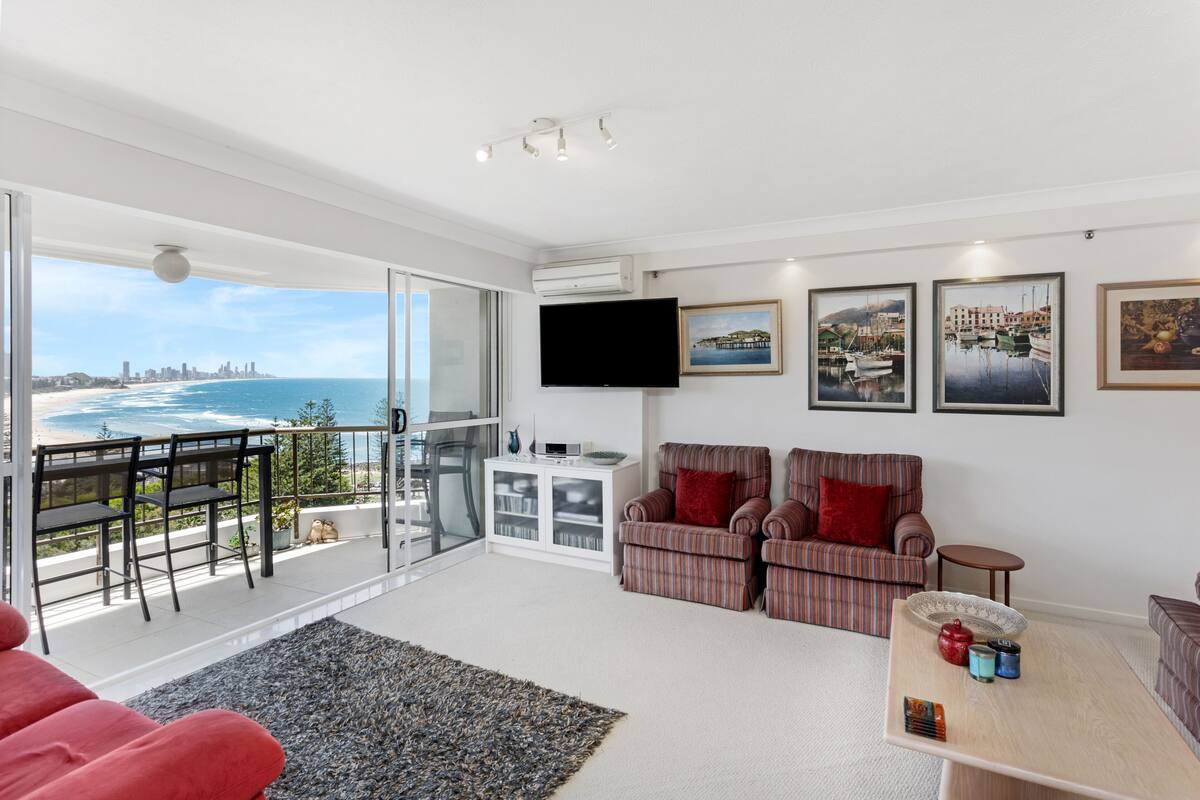 A modern living room with a large window overlooking a beach and cityscape, a flat-screen TV, a sofa, and a coffee table.