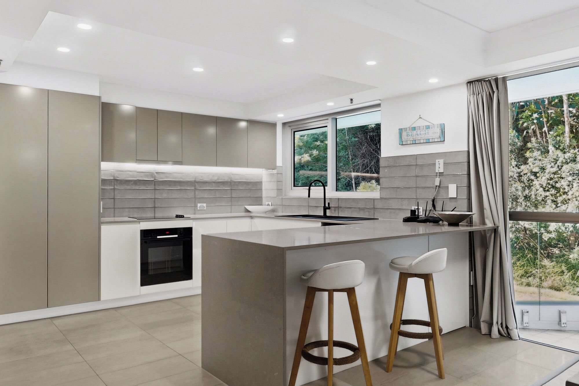 A modern kitchen with a central island, built-in oven, and a large window with a view of greenery.