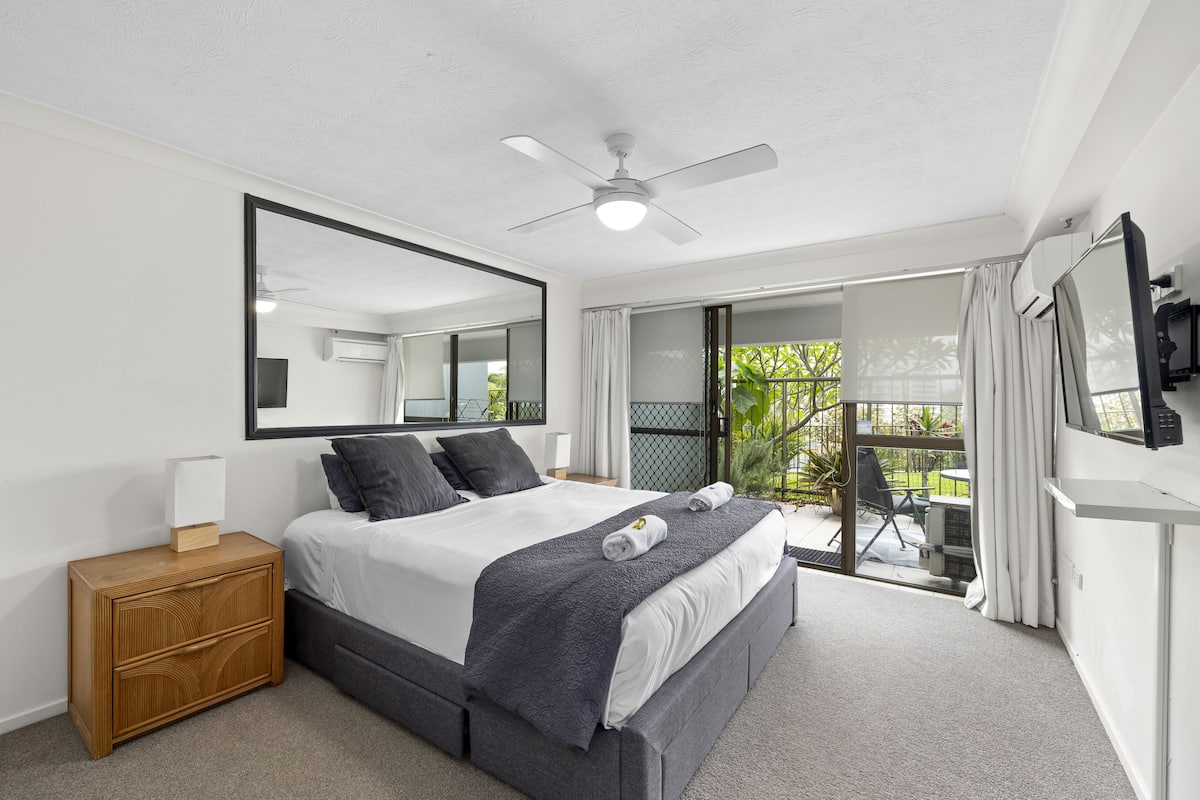 A bedroom with a bed, bedside table, ceiling fan, and a view of the outside balcony.