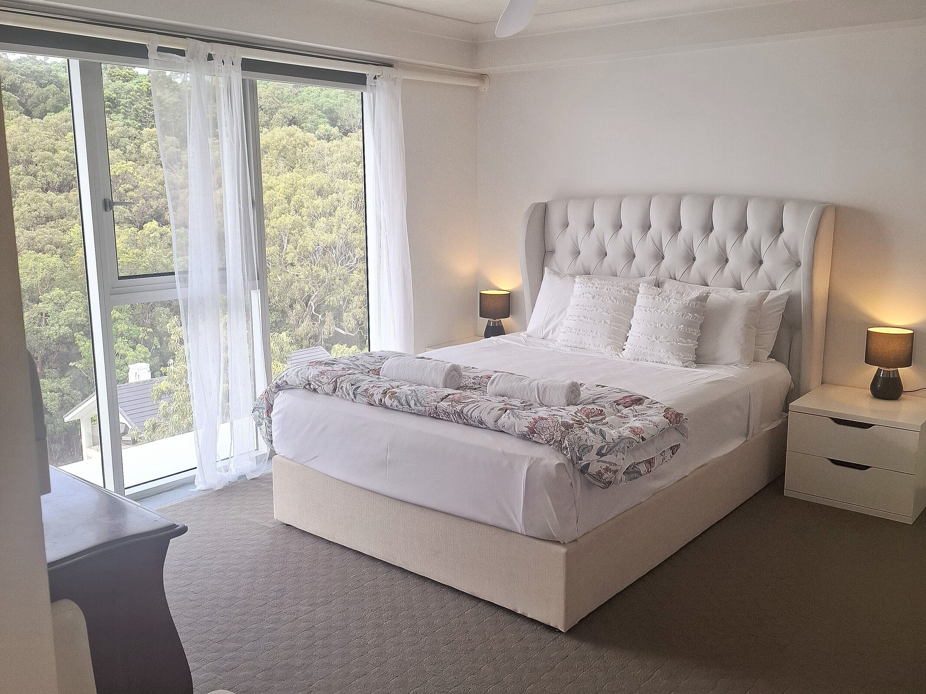 Quarto com cama grande, mesa de cabeceira, abajur e vista para árvores pela janela.