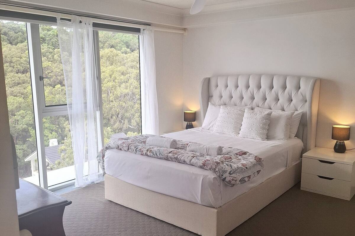 A bedroom with a large bed, a nightstand, a lamp, and a view of trees through a window.