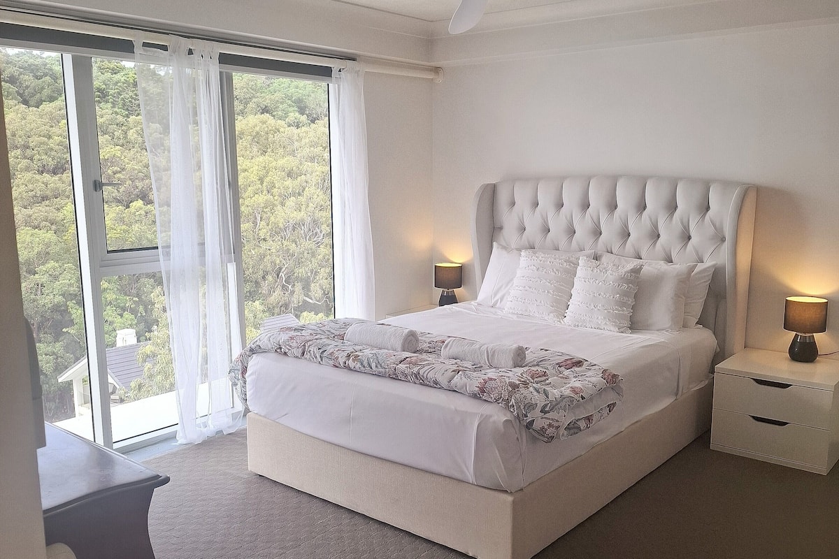 A bedroom with a large bed, a nightstand, a lamp, and a view of trees through a window.