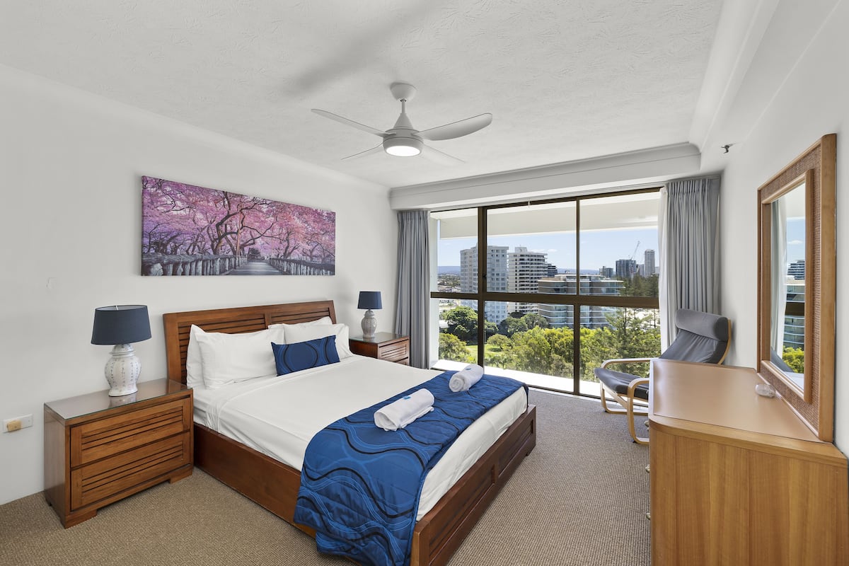 A hotel room with a large bed, a ceiling fan, a city view, and a desk with a chair.