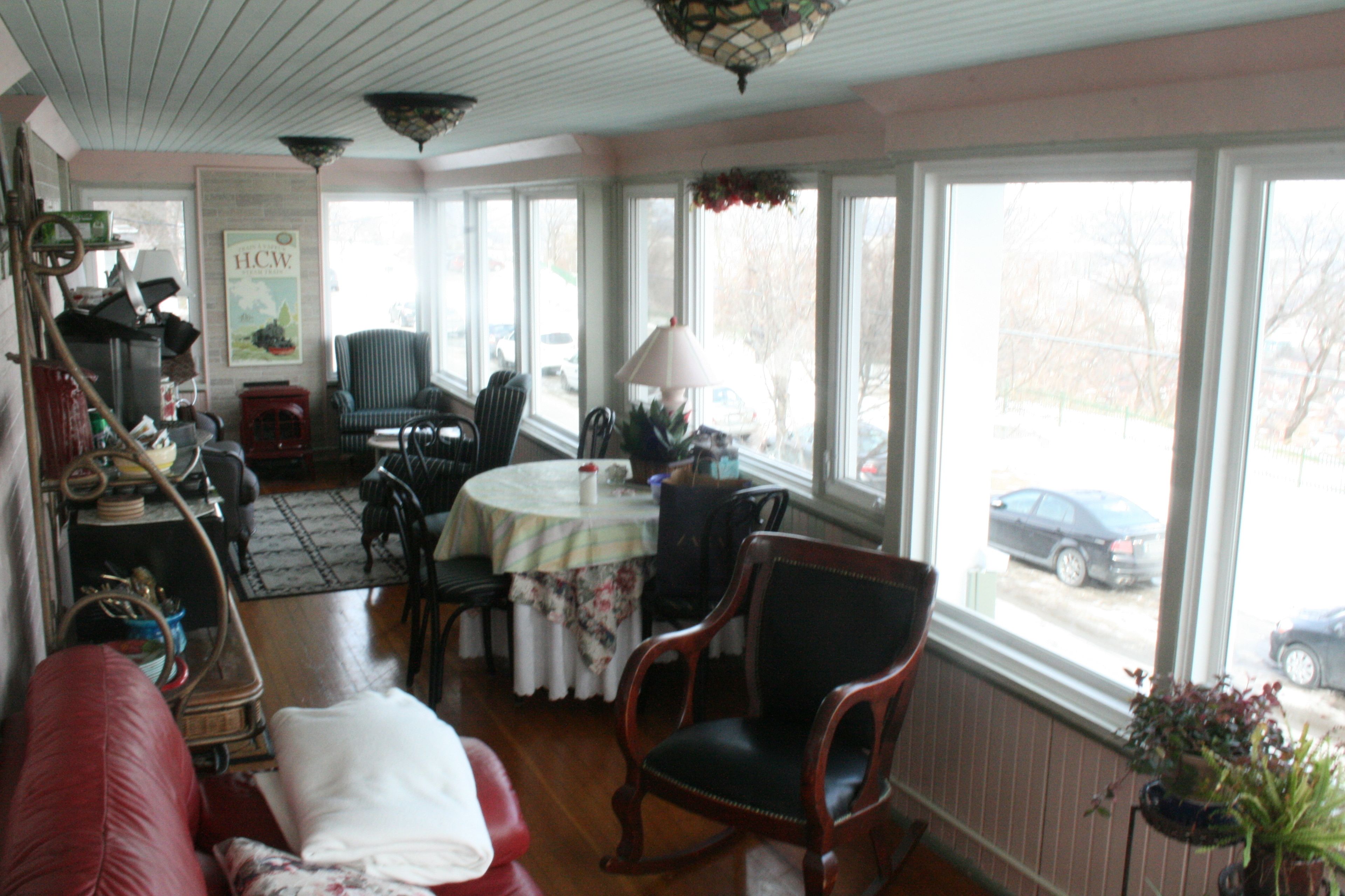 A cozy room with a red couch, a wooden rocking chair, a dining table with chairs, and large windows offering a view of the street.