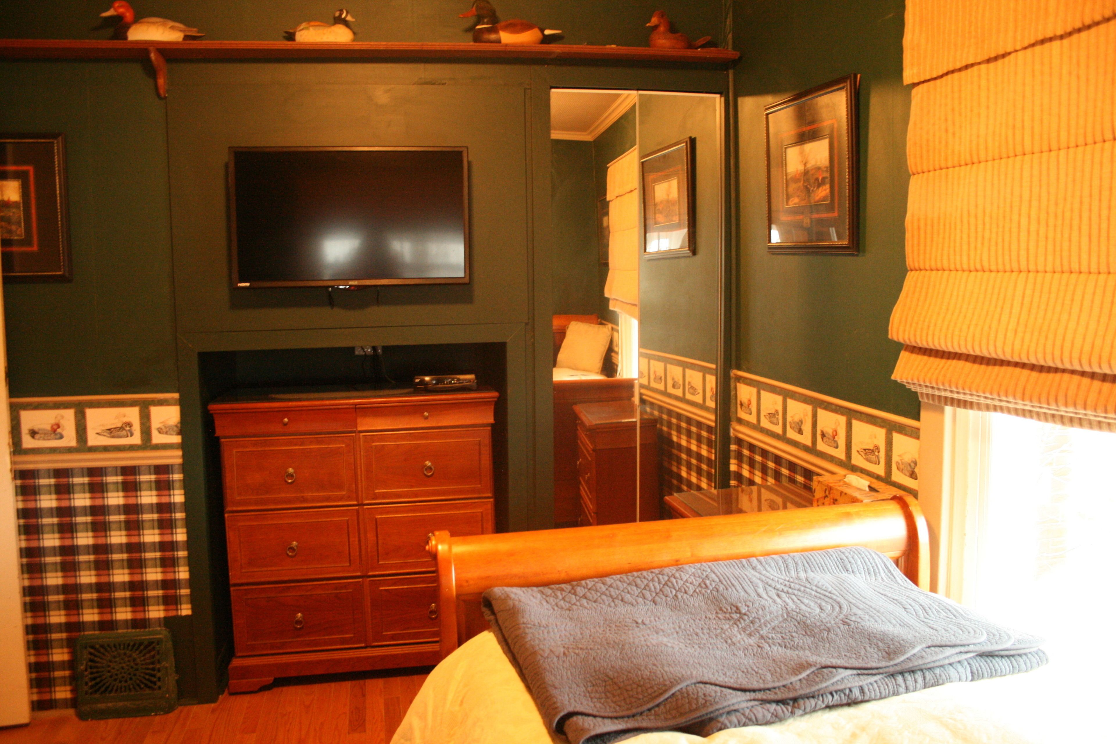 A room with a bed, a wooden dresser, a television mounted on the wall, and a window with blinds.
