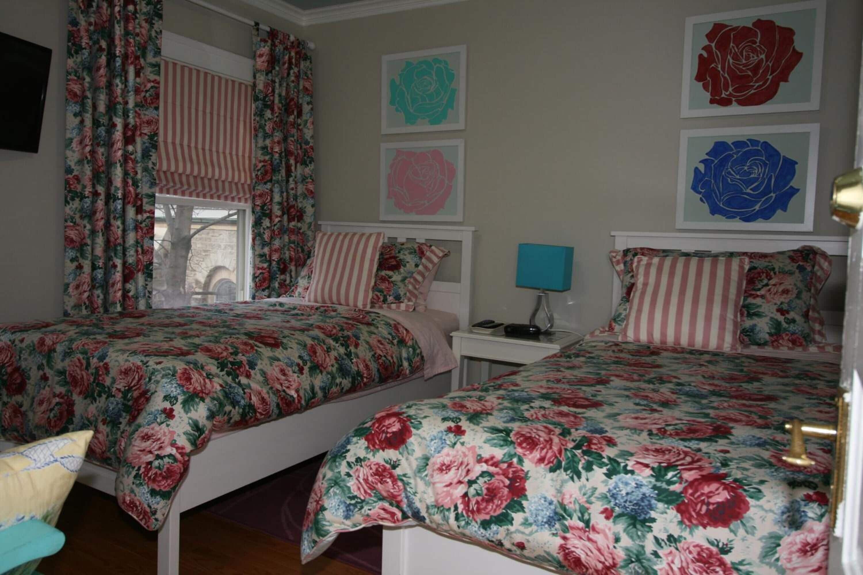 Une chambre avec deux lits, une literie à motifs floraux, une fenêtre avec des rideaux et des œuvres d’art murales.