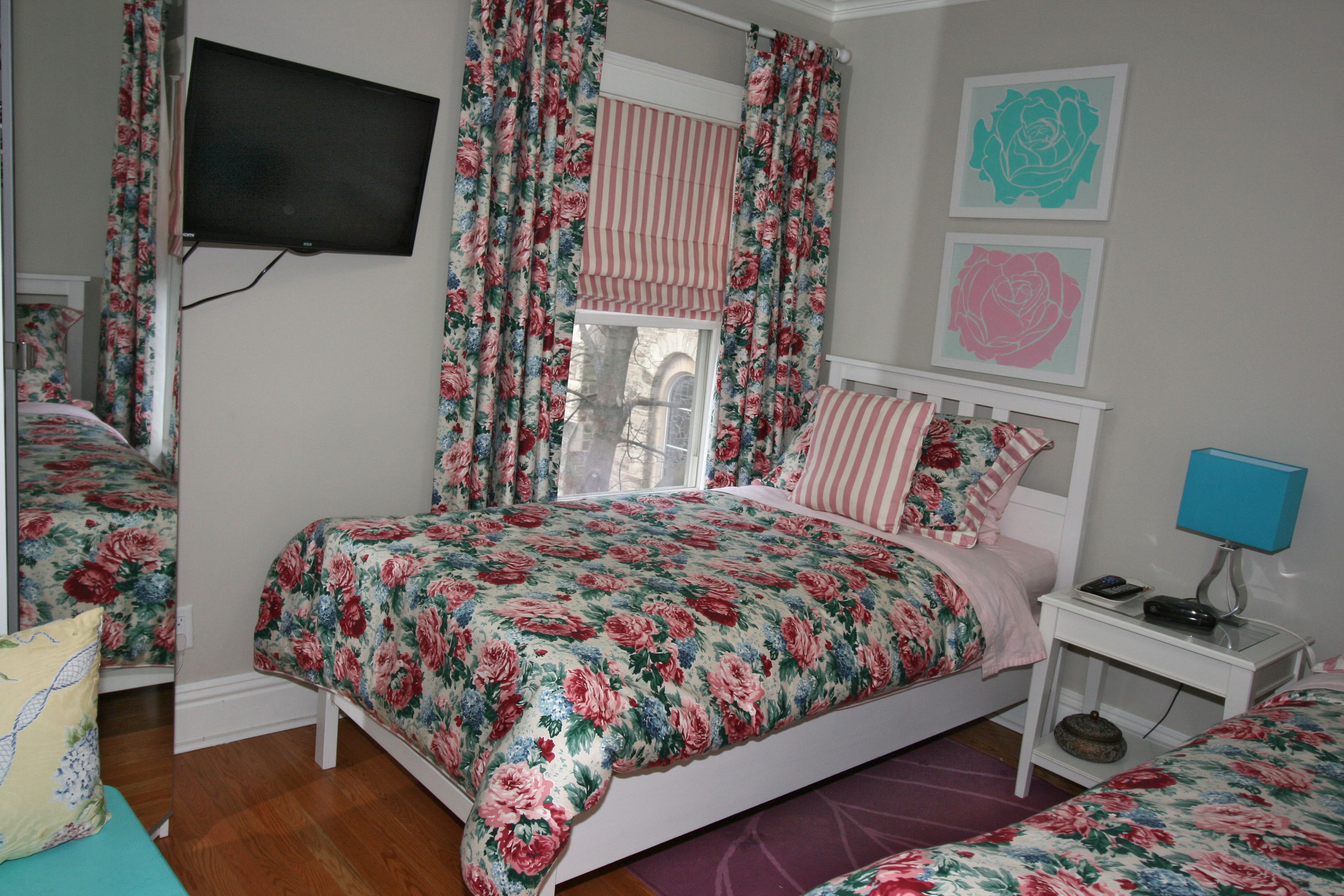 A bedroom with a floral bedspread, a television mounted on the wall, a window with curtains, and a bedside table with a lamp.