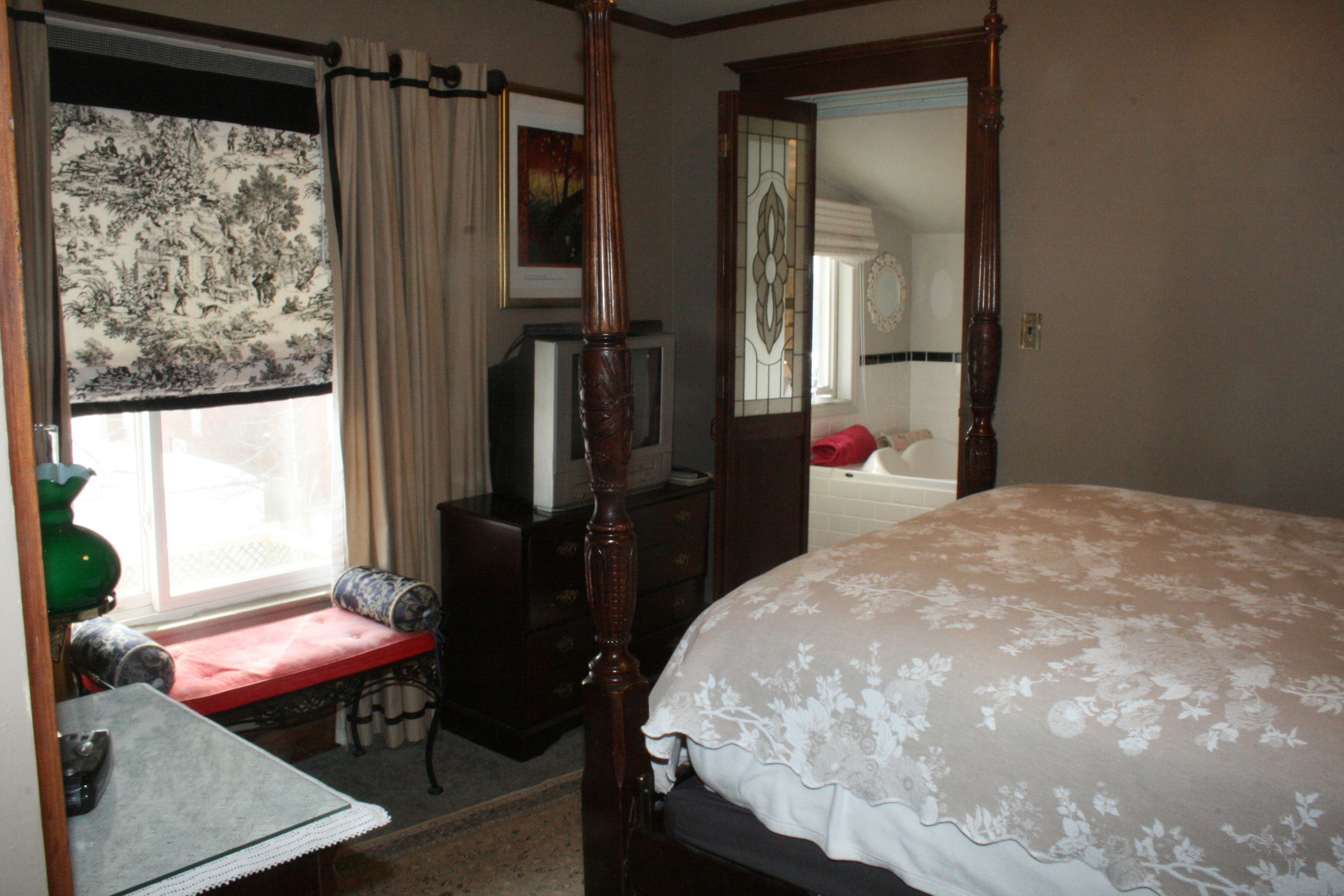 A bedroom with a four-poster bed, a vanity with a mirror, a dresser, a bench by the window, and a television on a stand.