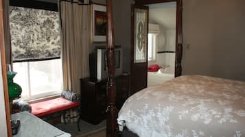 A bedroom with a four-poster bed, a vanity with a mirror, a dresser, a bench by the window, and a television on a stand.