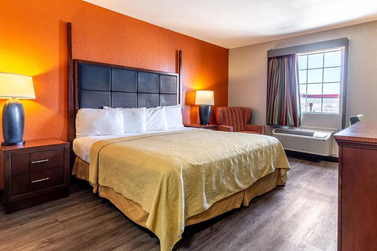 A hotel room with a large bed, two bedside tables with lamps, a chair, a window with curtains, and a radiator.