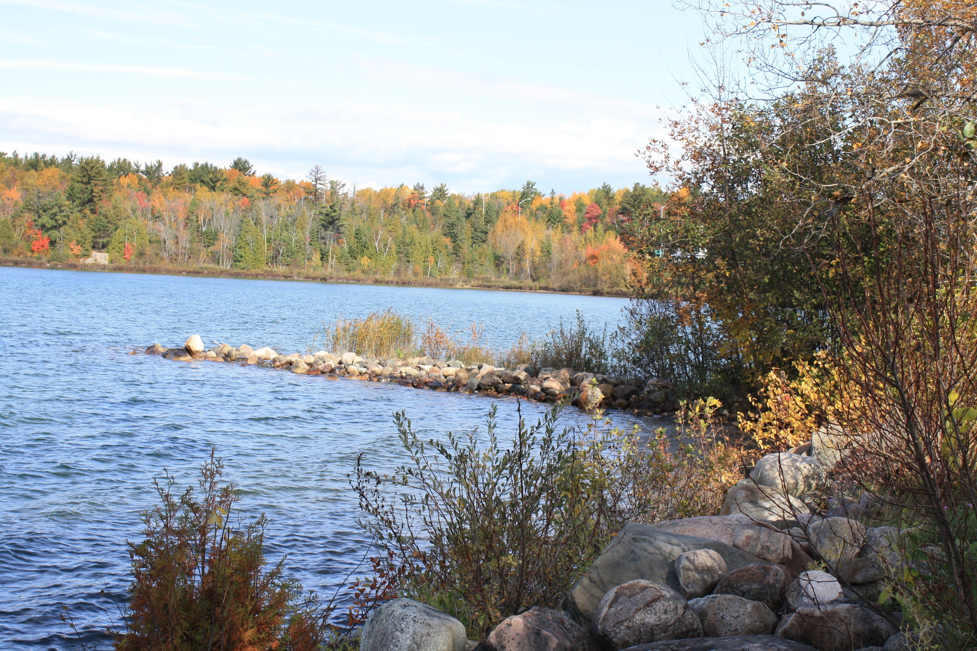 A calm lake with a rocky shoreline and autumn-colored trees in the background.