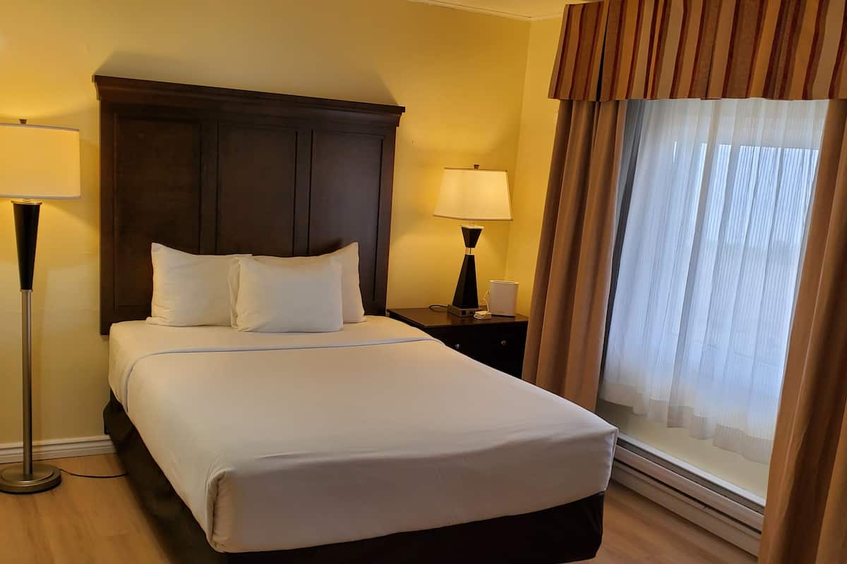 A hotel room with a bed, two bedside lamps, a nightstand, a window with curtains, and a wooden floor.