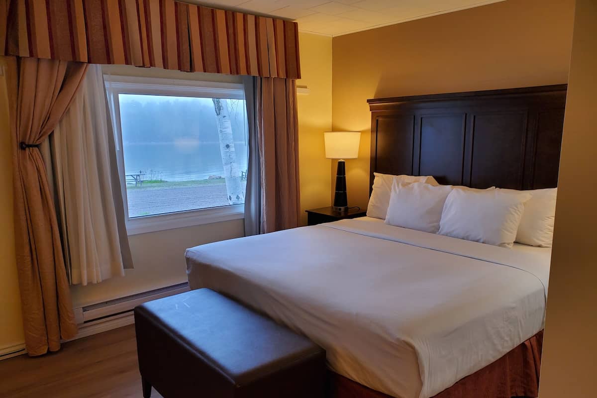 A hotel room with a large bed, a bedside table with a lamp, a window with curtains, and a view of the beach.