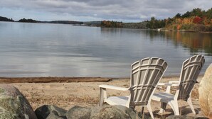 Dos sillas Adirondack sobre una orilla arenosa junto a un lago tranquilo, con follaje otoñal al fondo.