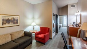 A hotel room with a sofa, a red armchair, a desk with a lamp, a framed picture on the wall, and a hallway leading to a bathroom. - Country Inn & Suites by Radisson, El Dorado, AR (El Dorado)