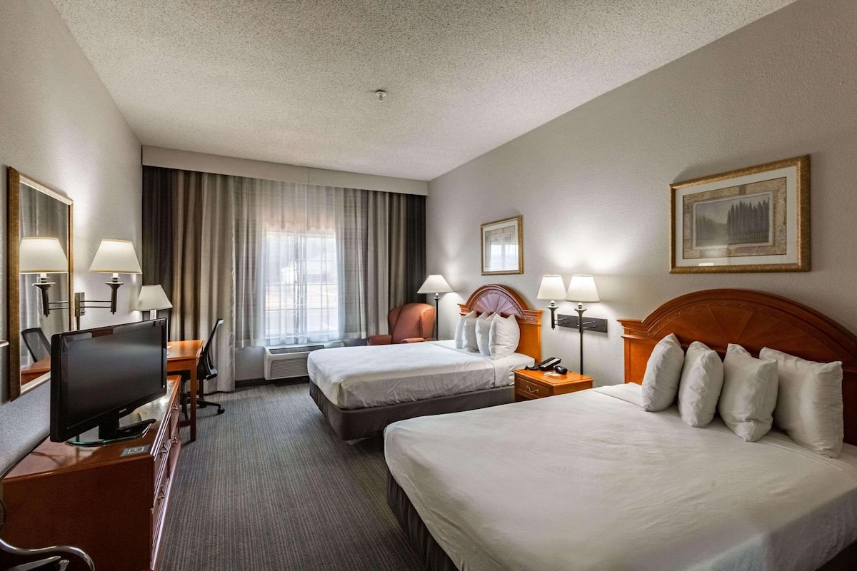 A hotel room with two beds, a desk with a television, a chair, a window with curtains, and a framed picture on the wall.