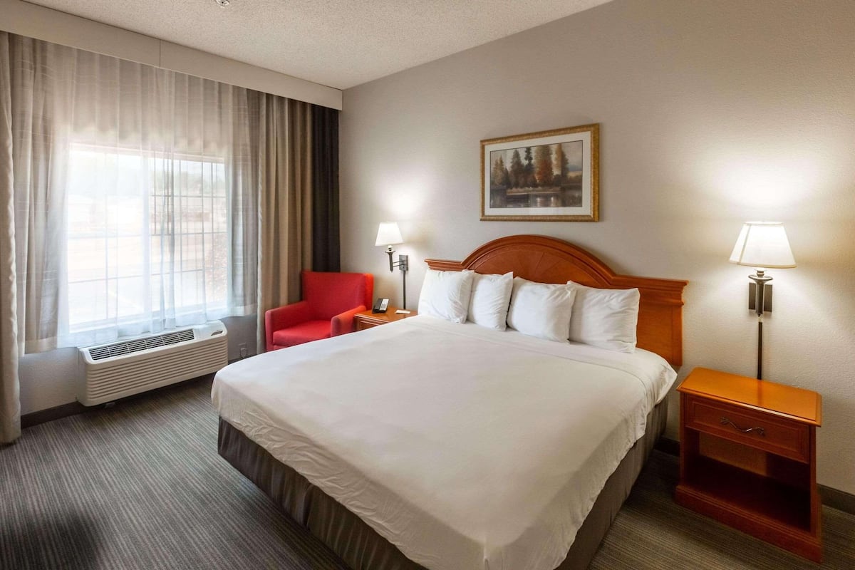 A hotel room with a large bed, a red chair, two bedside tables with lamps, a painting on the wall, and a window with curtains.