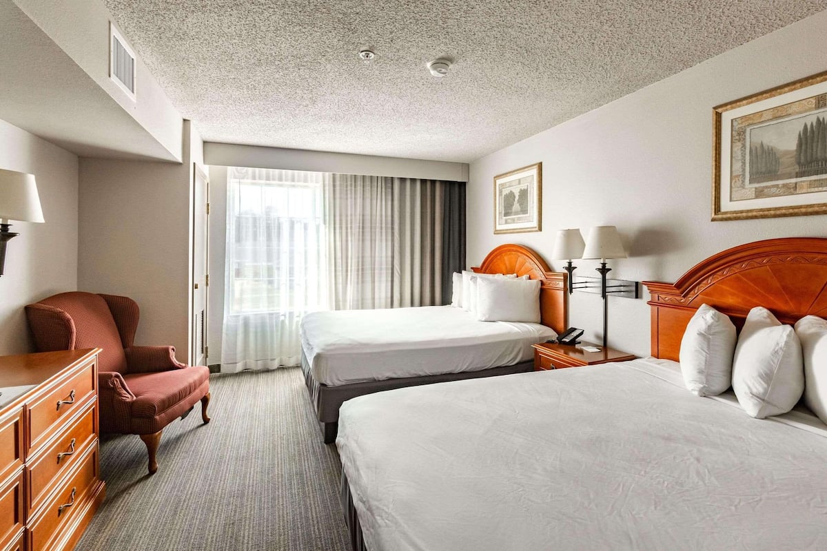 A hotel room with two beds, a wooden dresser, a chair, and a large window with curtains.