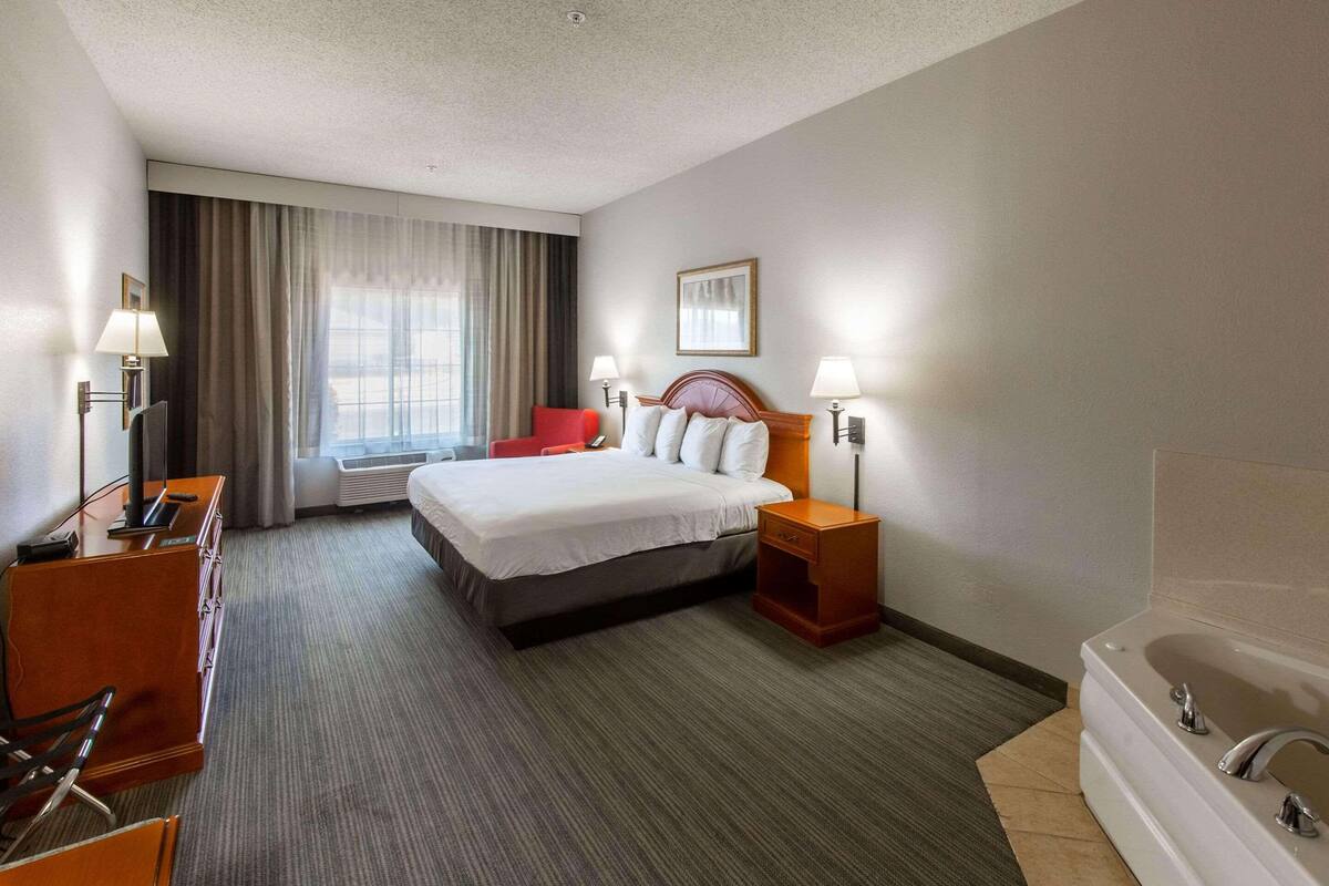 A hotel room with a bed, a desk, a chair, a red chair, a nightstand with a lamp, a bathroom with a bathtub, and a window with curtains.
