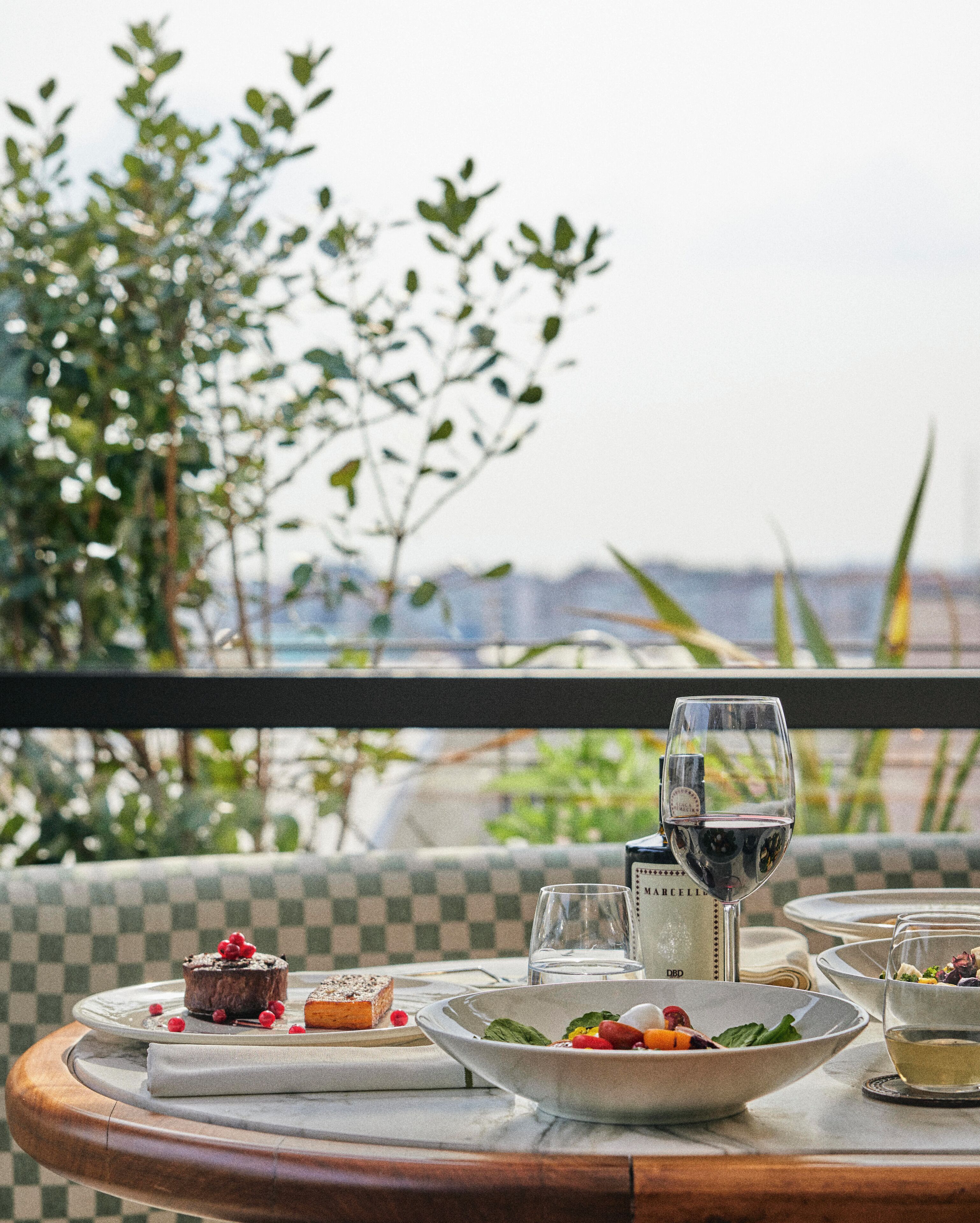A dining table with a checkered tablecloth, a dessert plate with berries, a wine glass, and a bowl of salad.