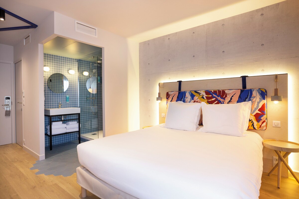 Une chambre d’hôtel moderne avec un grand lit, une salle de bain avec un mur recouvert de carreaux bleus et une peinture abstraite aux couleurs vives accrochée au mur.