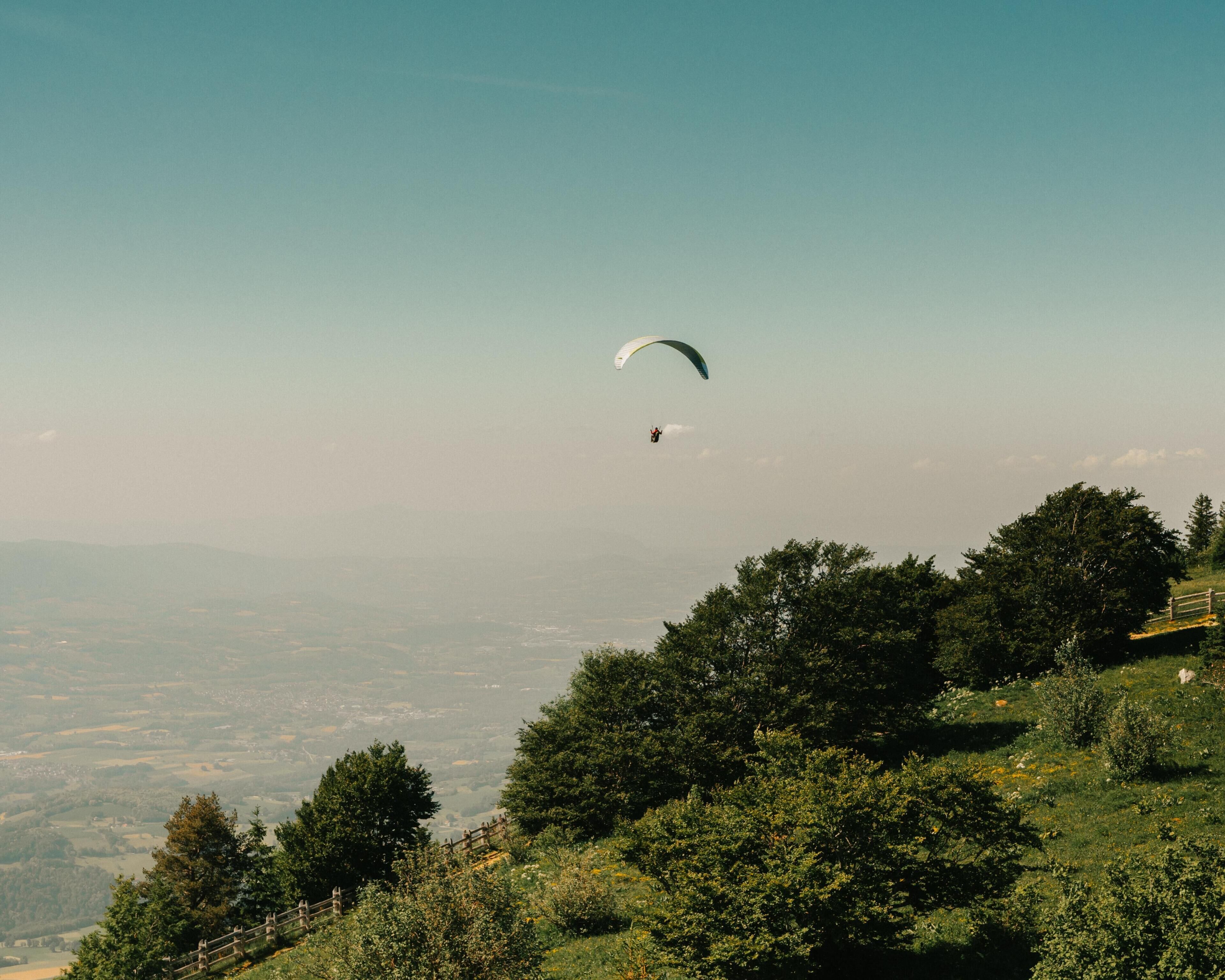 Una persona practica parapente sobre un paisaje con árboles y una vista distante de un pueblo.