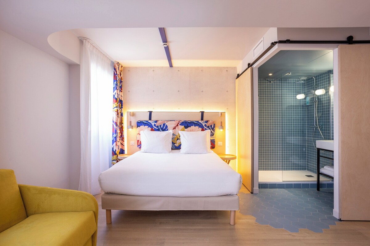 Une chambre d’hôtel moderne avec un grand lit, un fauteuil jaune, un espace douche et une salle de bain équipée d’un lavabo et d’un miroir.