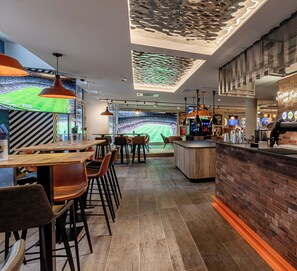 A modern bar area with wooden tables and chairs, a brick accent wall, and a large screen displaying a sports event.
