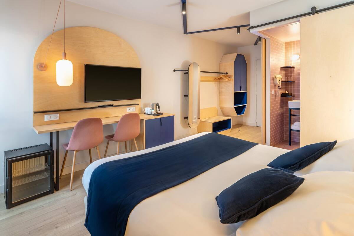 Une chambre d’hôtel moderne équipée d’un lit, d’un bureau, de chaises, d’une télévision et d’une salle de bain visible à travers une porte ouverte.
