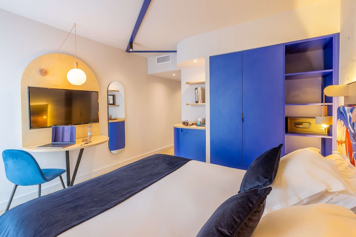 Une chambre d’hôtel moderne équipée d’un lit, d’un bureau avec un ordinateur portable, d’une télévision et d’une armoire bleue.