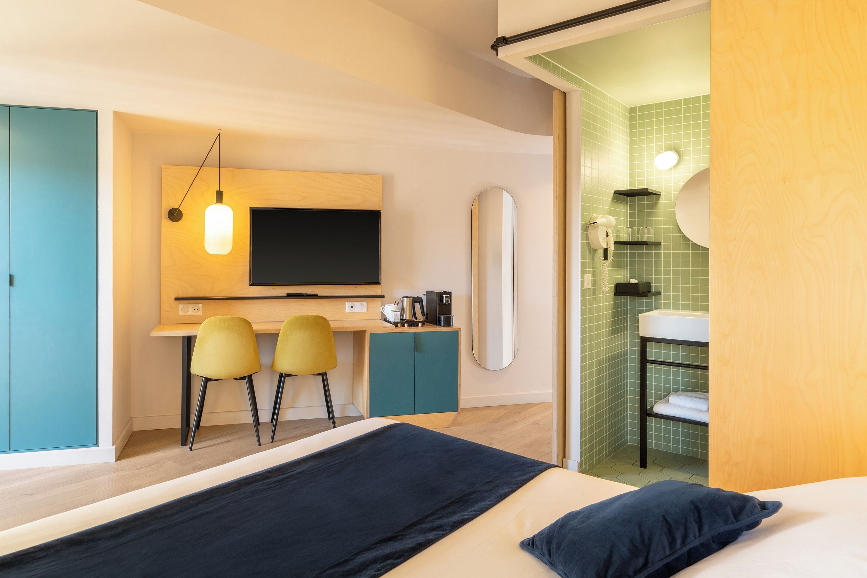 A modern hotel room with a bed, a desk with two chairs, a television, and a bathroom with a sink and mirror.