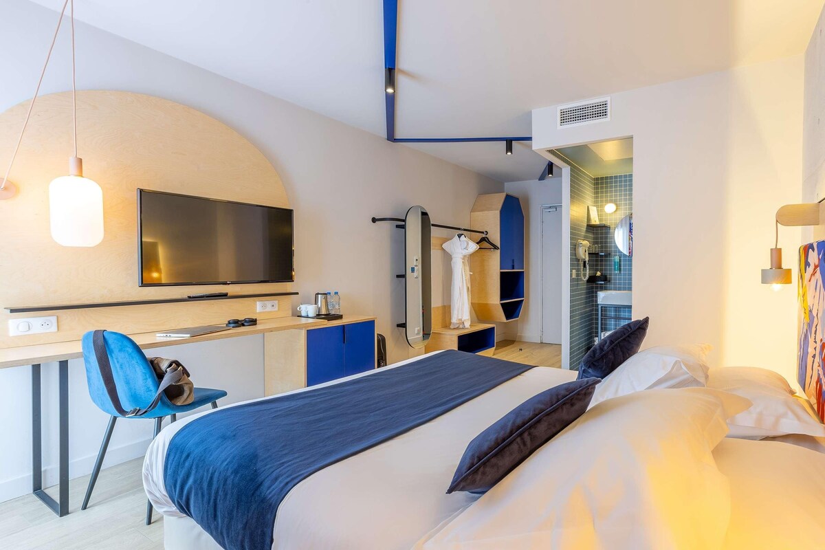 Une chambre d’hôtel moderne équipée d’un lit, d’un bureau, d’une chaise, d’une télévision et d’une salle de bain visible à travers une porte ouverte.