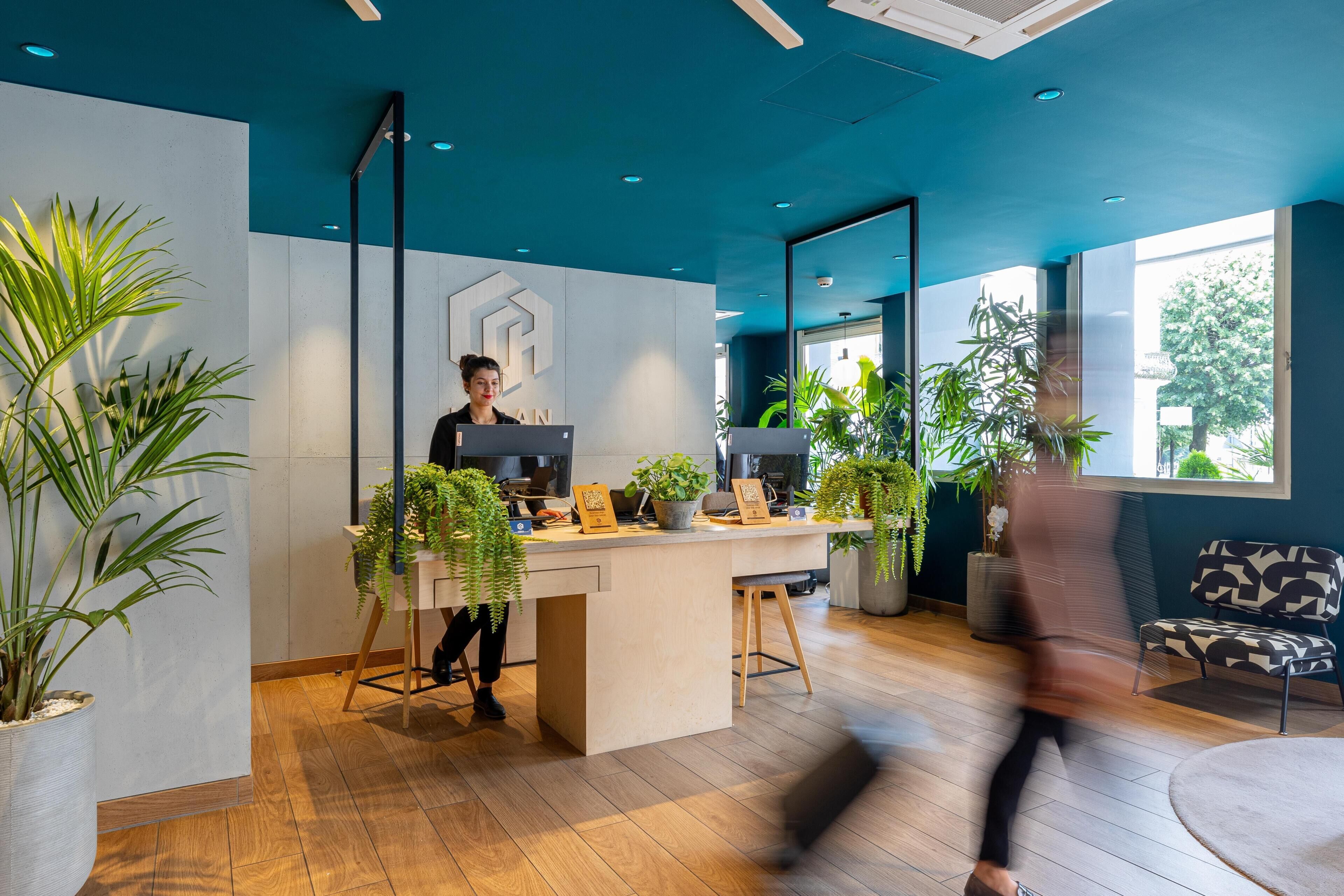 Un espacio de oficina moderno con un mostrador de recepción, una persona sentada frente a una computadora y varias plantas en macetas.