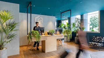 A modern office space with a reception desk, a person seated at a computer, and various potted plants.