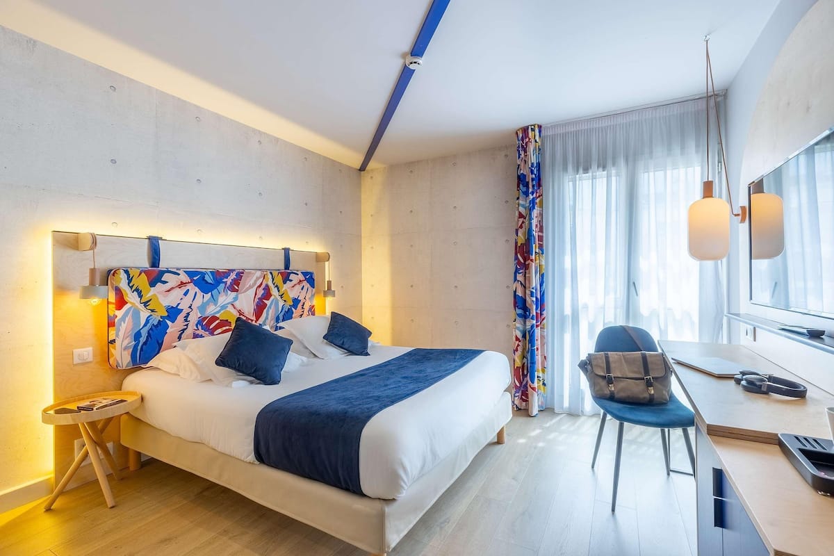 Une chambre d’hôtel moderne équipée d’un lit, d’un bureau, d’une chaise et d’une grande fenêtre avec des rideaux.