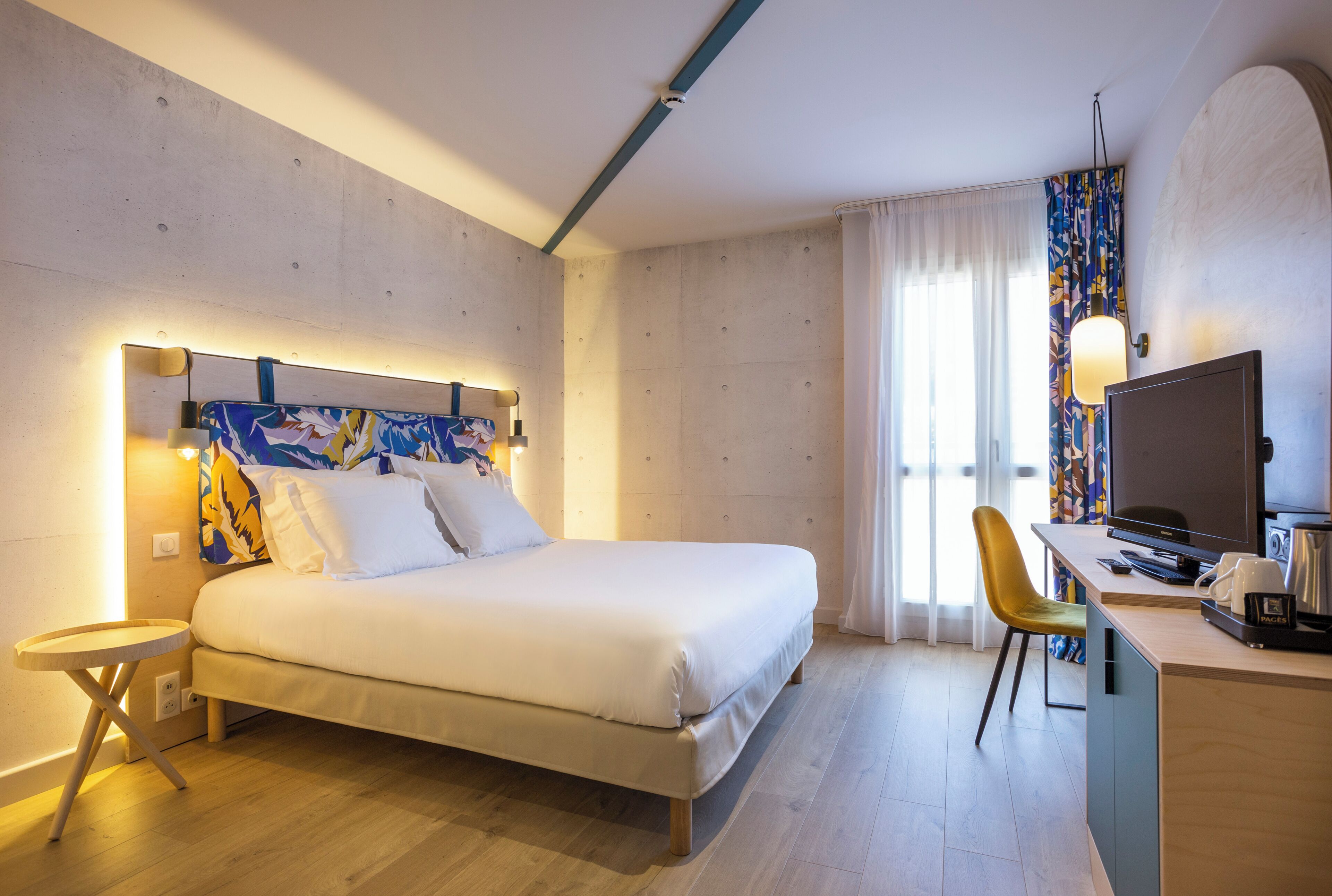A modern hotel room with a bed, a desk, a television, and a window with curtains.