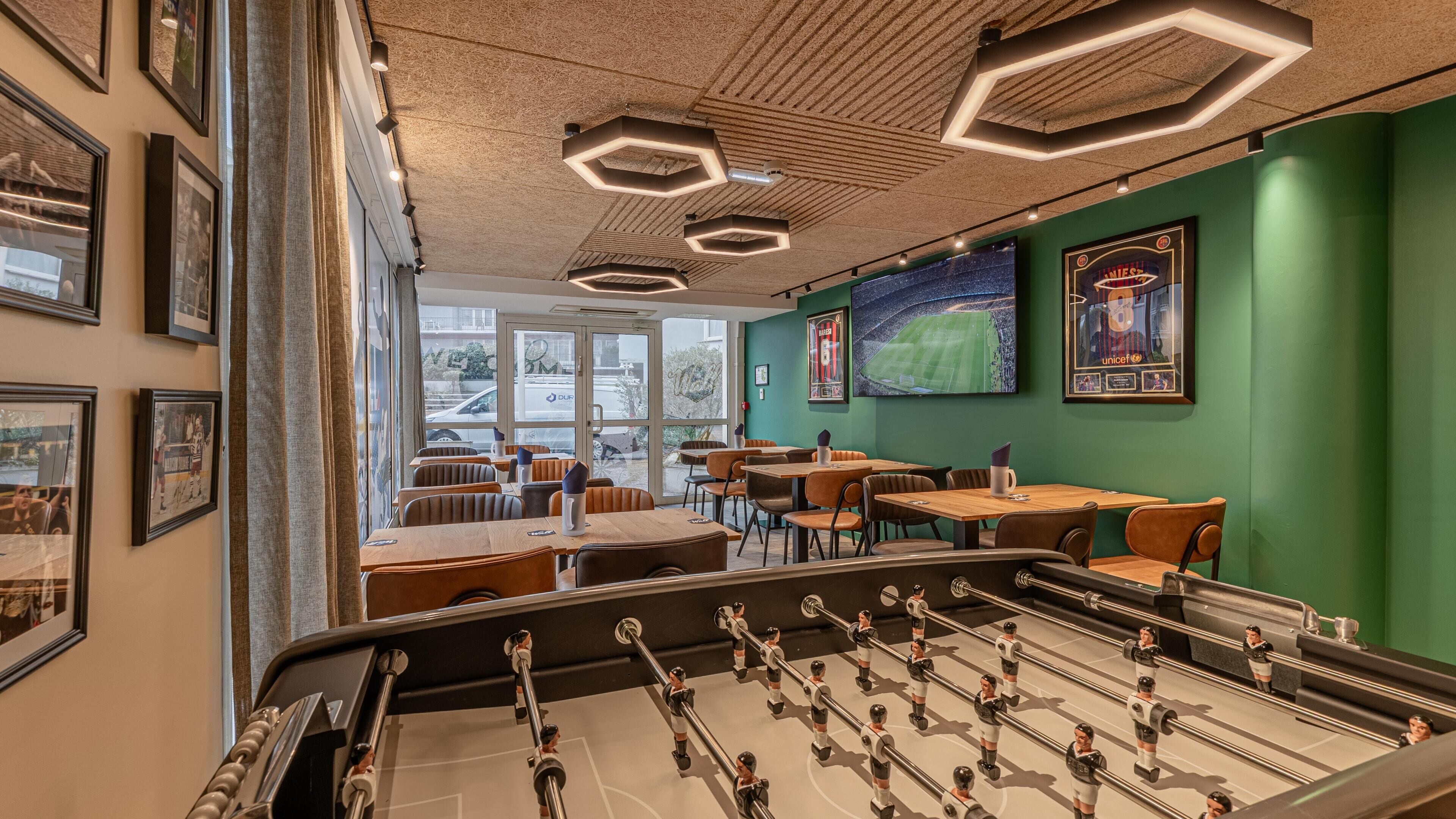 A room with a foosball table, framed sports memorabilia on the walls, and a view of the outdoors through large windows.