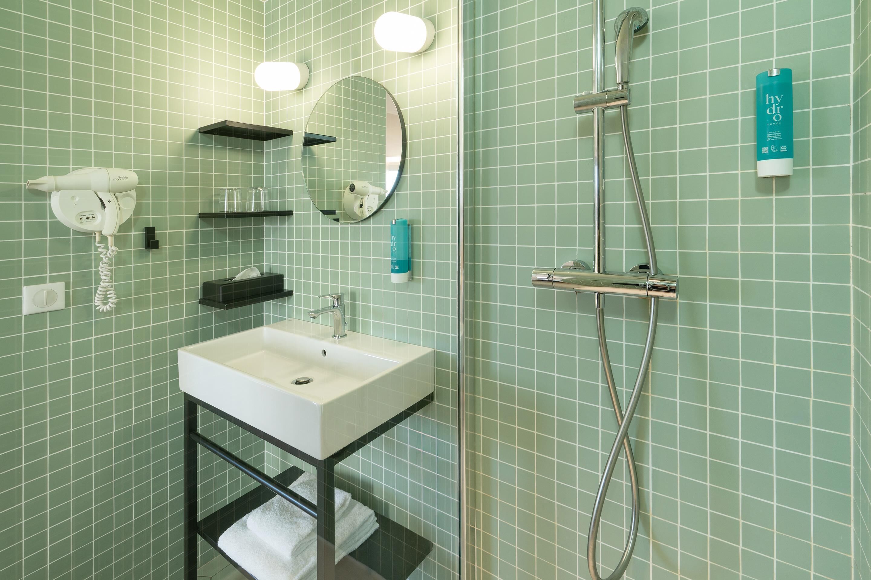 Un baño con lavamanos blanco, espejo, secador de pelo, toallero y ducha con cabezal de ducha de mano.