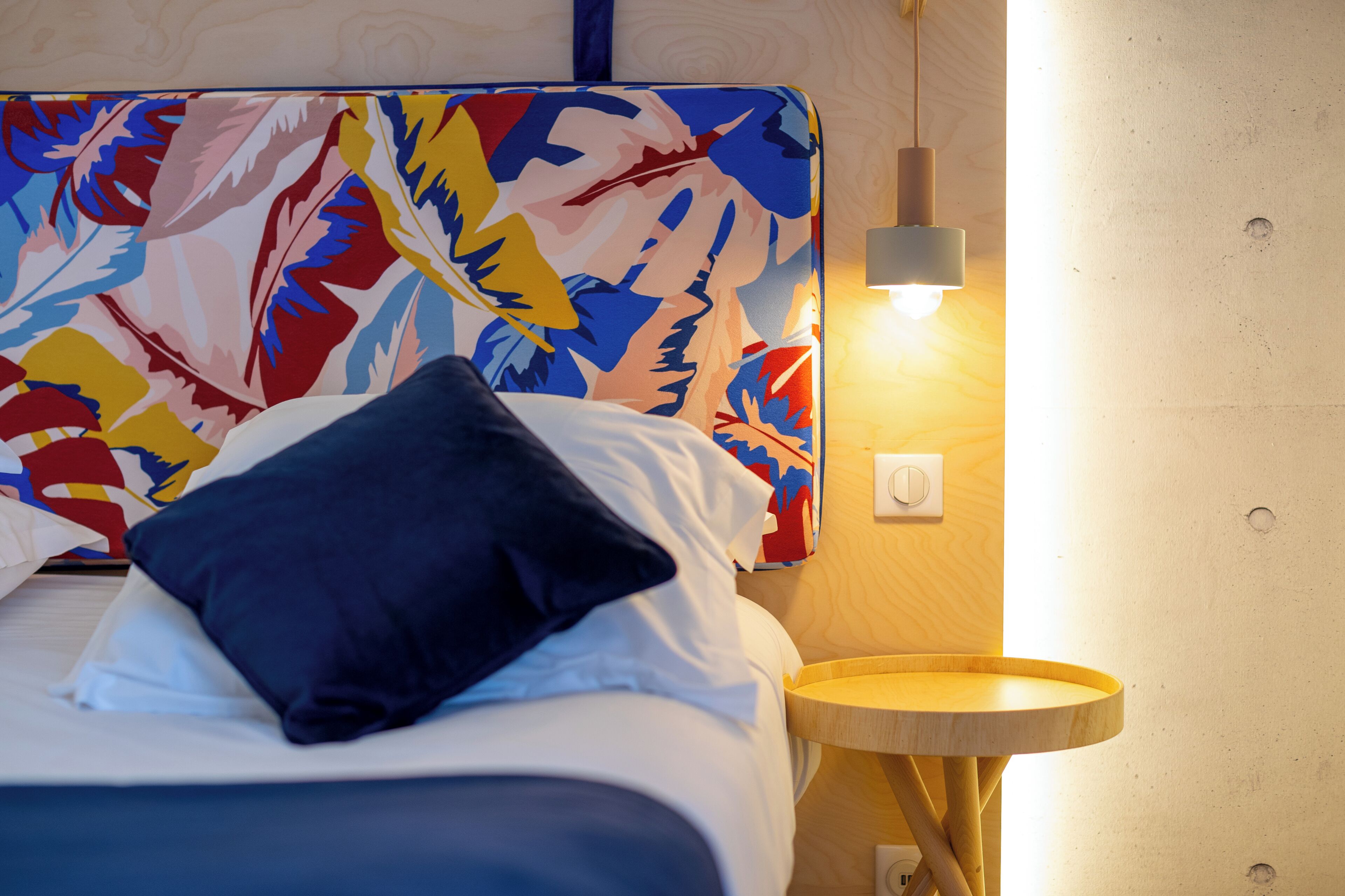 A bed with a colorful headboard, a bedside lamp, and a round wooden nightstand.