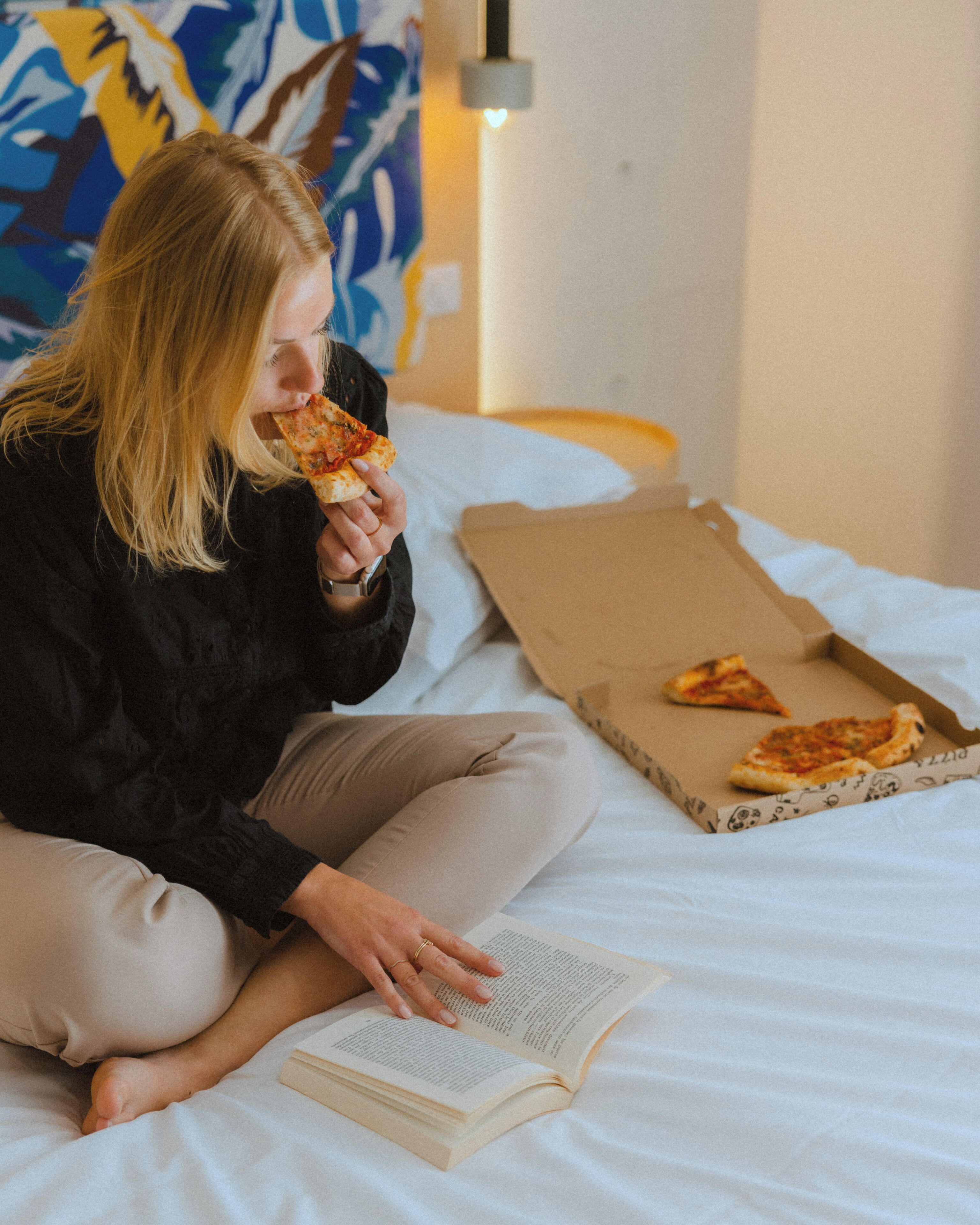 A person sitting on a bed with a pizza box and a book.