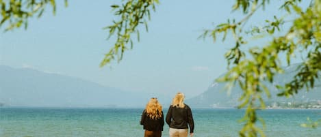 Dos personas caminando por una playa, con agua y montañas al fondo.