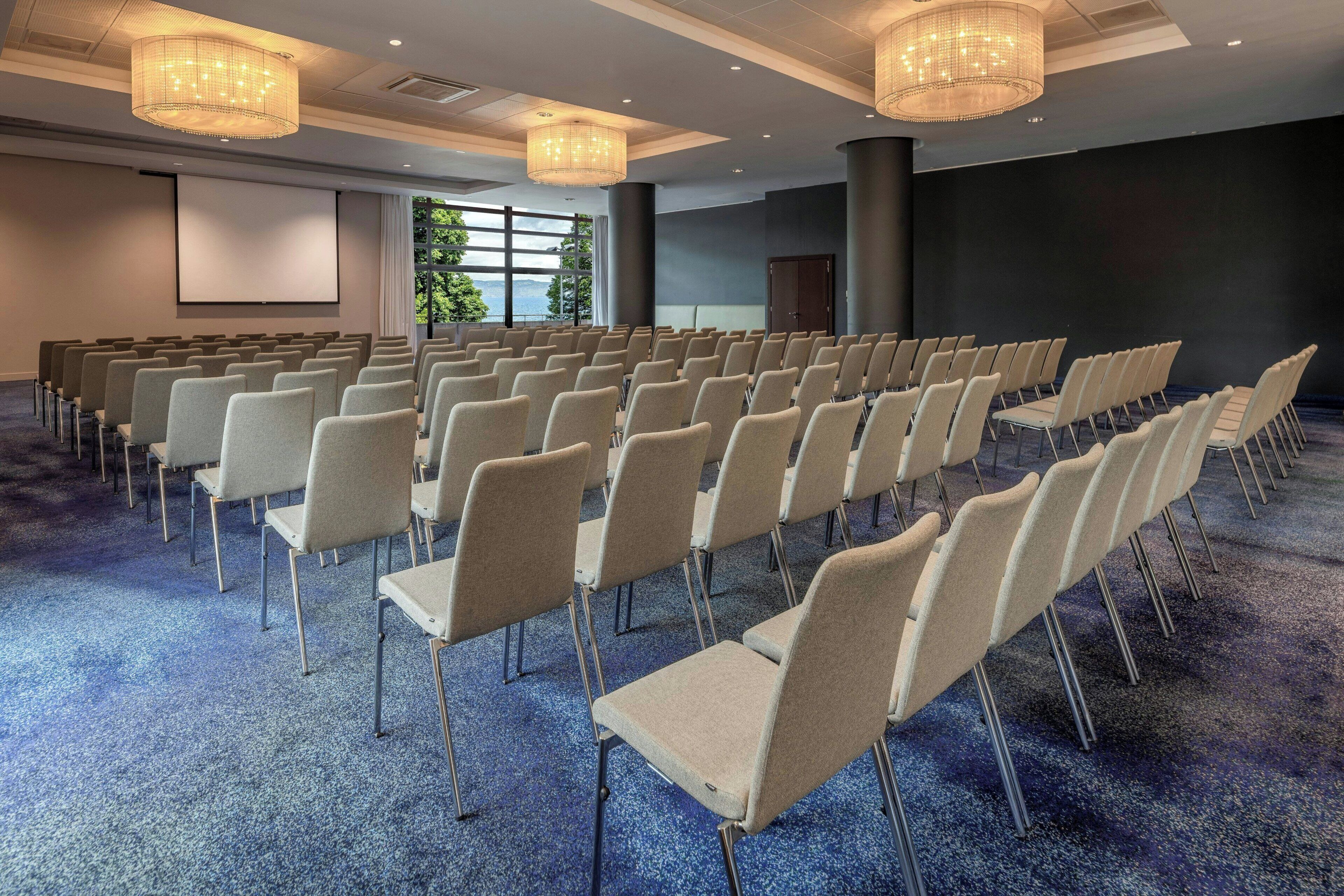 Un salón de conferencias con filas de sillas, una pantalla de proyector y amplias ventanas.