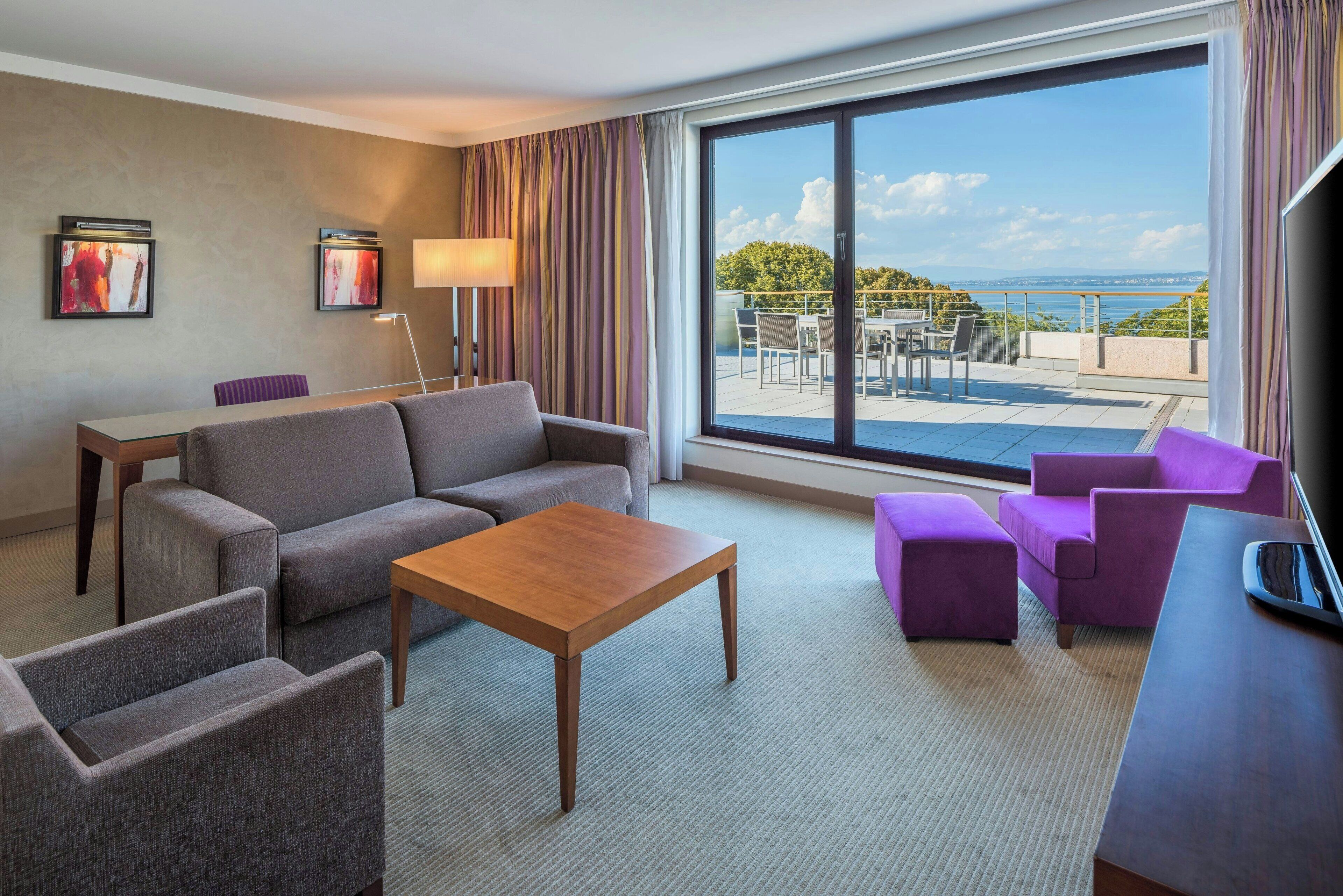 Habitación de hotel con sofá, sillas, una mesita de centro, escritorio y un ventanal con vistas al mar.